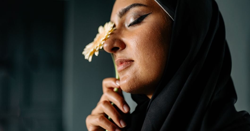 Woman in a grey abaya standing beside a window in soft indoor light, illustrating a calm, considered approach to abaya care