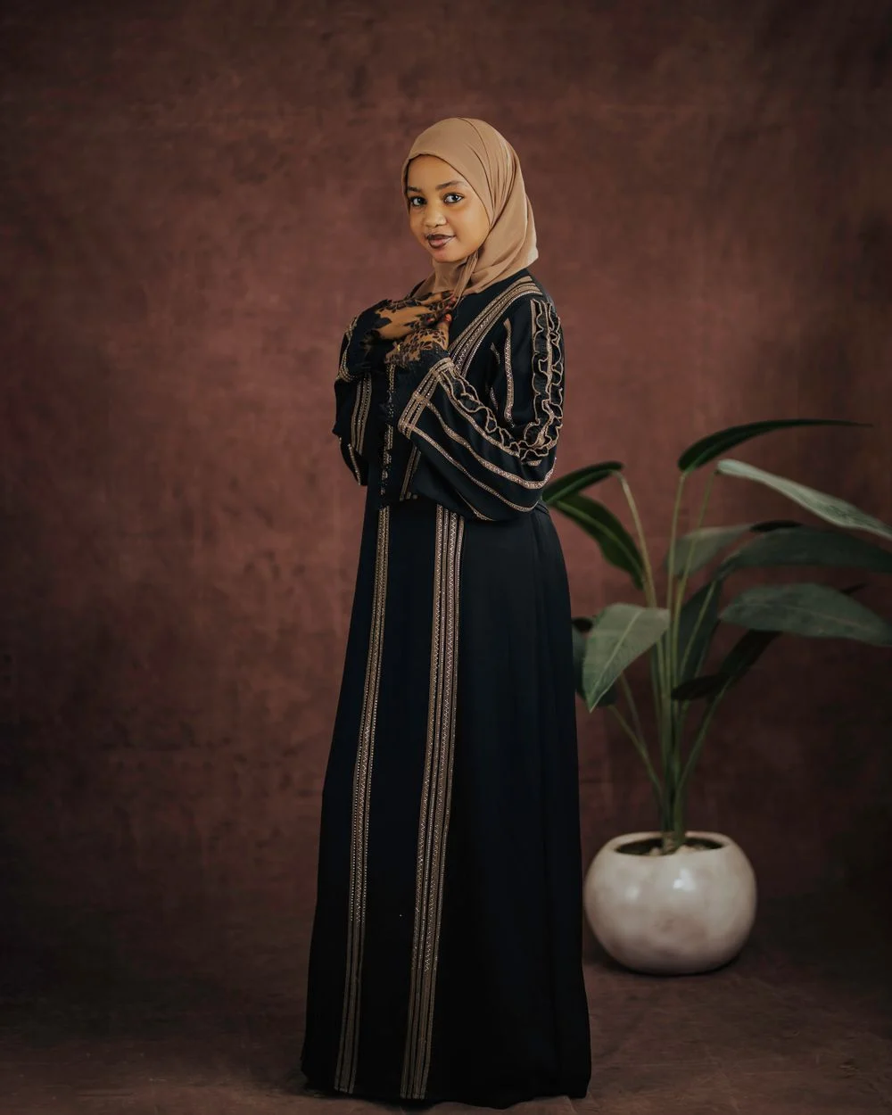 Woman in a black abaya with gold trim standing beside a plant against a brown studio backdrop