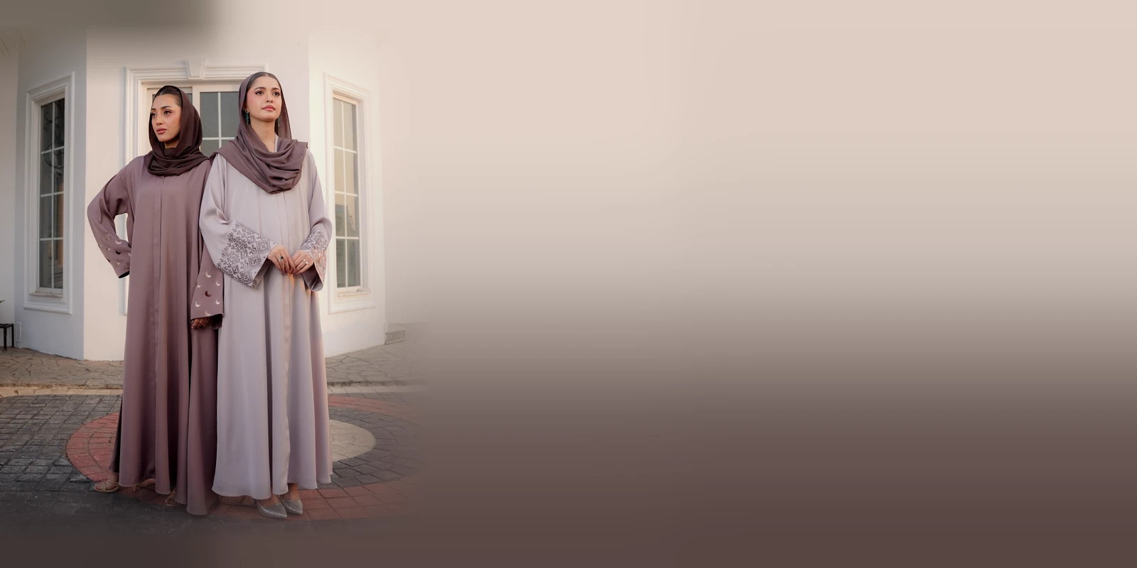 Two women in long abayas in dusty mauve and soft grey with matching headscarves, standing together in front of a white building with tall windows