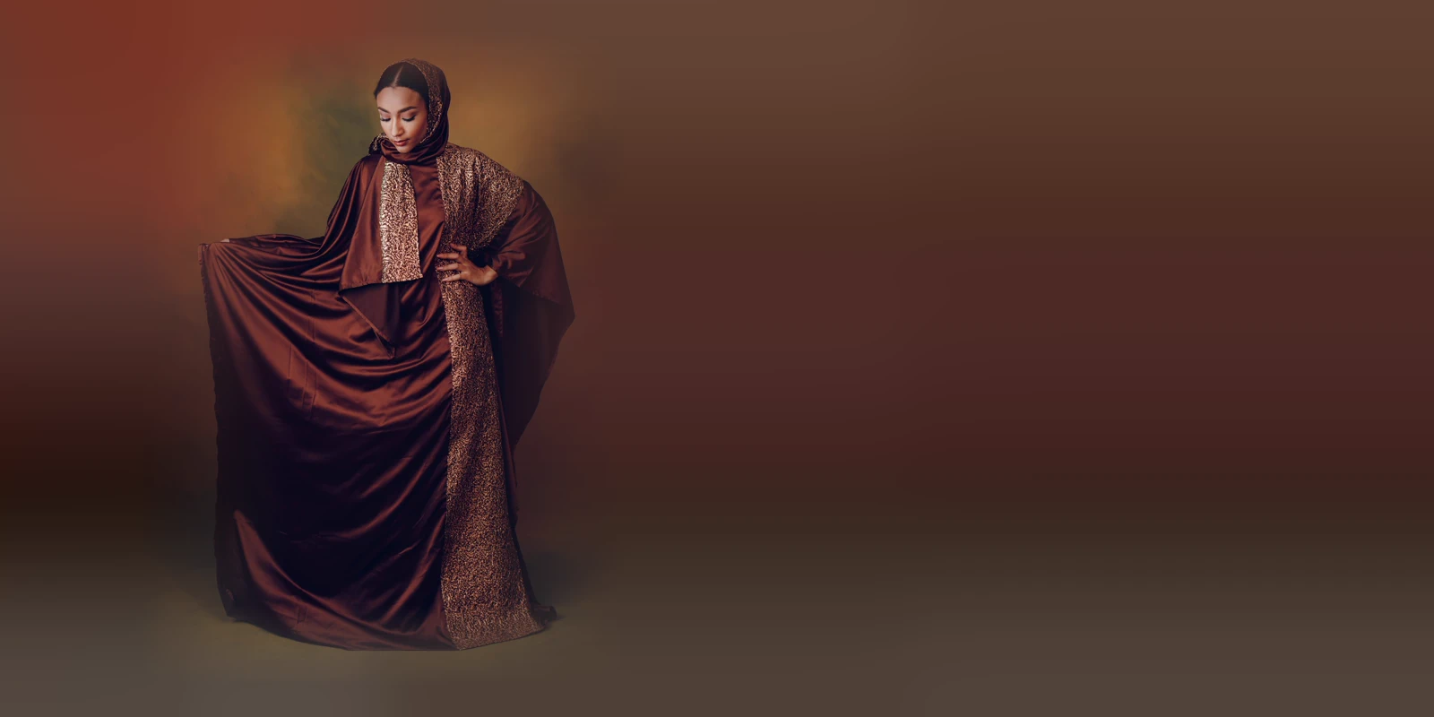 Woman in a chocolate-brown satin batwing abaya with a gold brocade panel and matching headscarf, holding out the fabric against a warm painterly backdrop