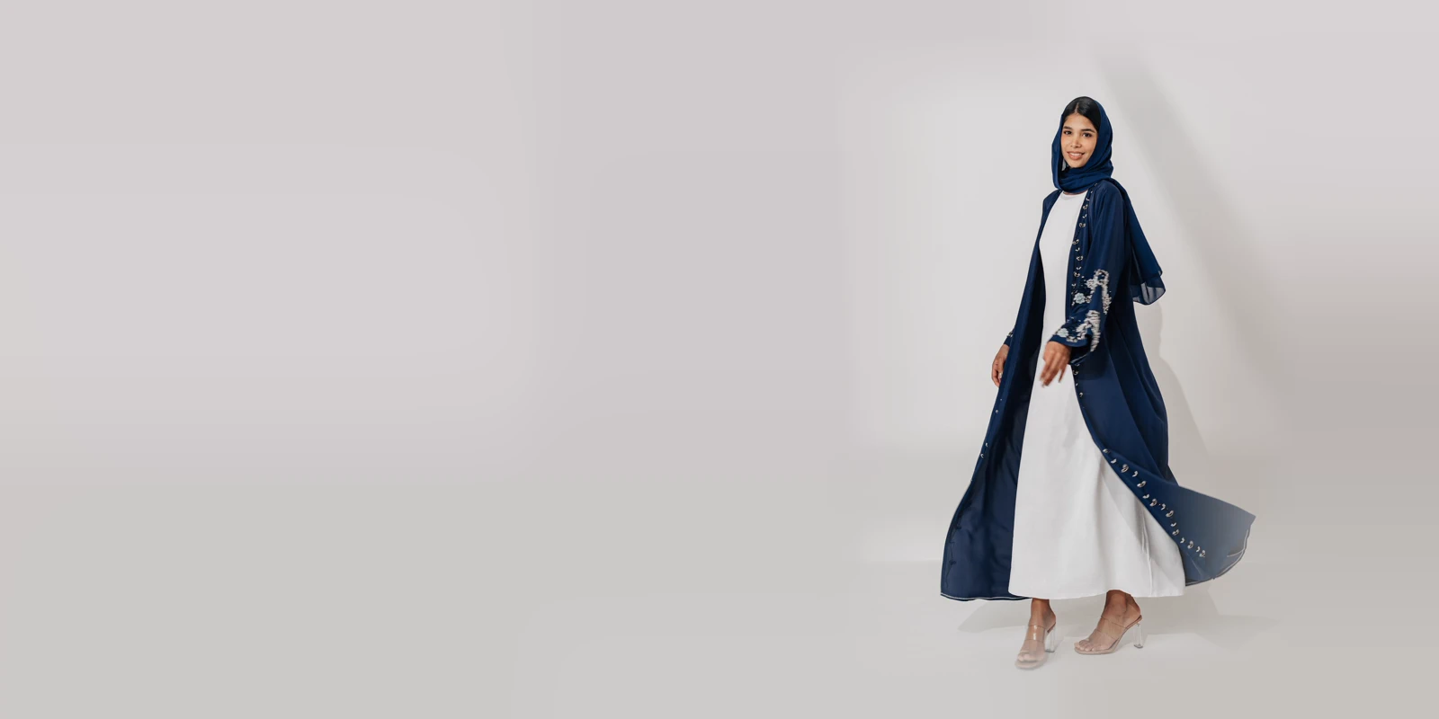 Woman in a navy open abaya with silver embroidery over a white dress and a navy headscarf, walking in a bright studio with soft window shadows