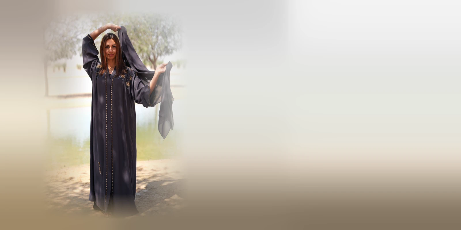 Woman in a charcoal open abaya with beaded trim lifting her sheer headscarf above her head beside an oasis pool with trees