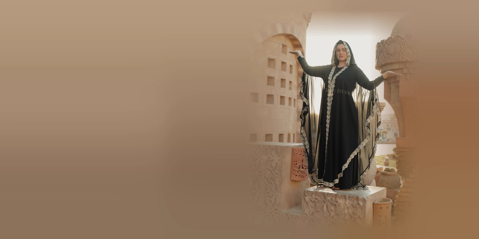 Woman in a black abaya with sheer batwing sleeves and cream lace trim standing with her arms out on carved sandstone steps, with ornate towers behind