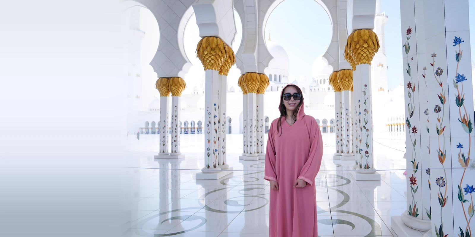 Woman in a long pink hooded abaya and sunglasses standing between white marble columns with floral inlay in a sunlit arcade