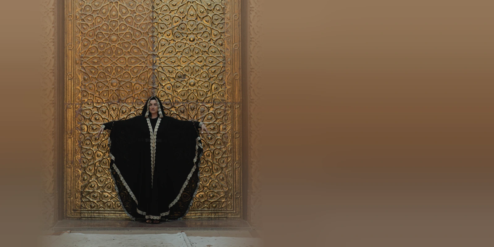 Woman in a black batwing abaya with a cream lace border standing with her arms outstretched in front of an ornate gold carved door