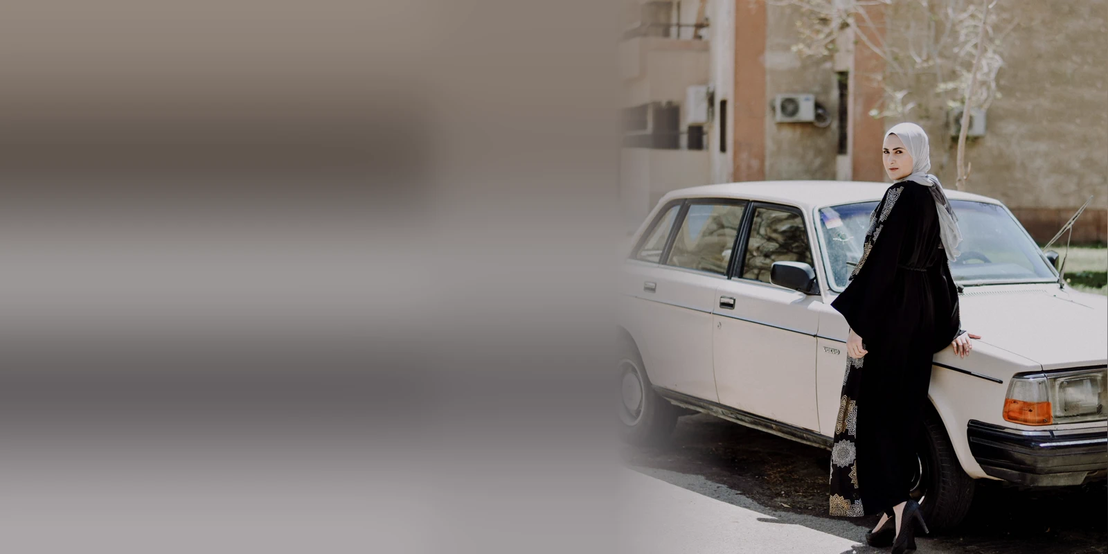 Woman in a black open abaya with embroidered trim and a pale headscarf leaning against a vintage cream saloon car on a sunlit street