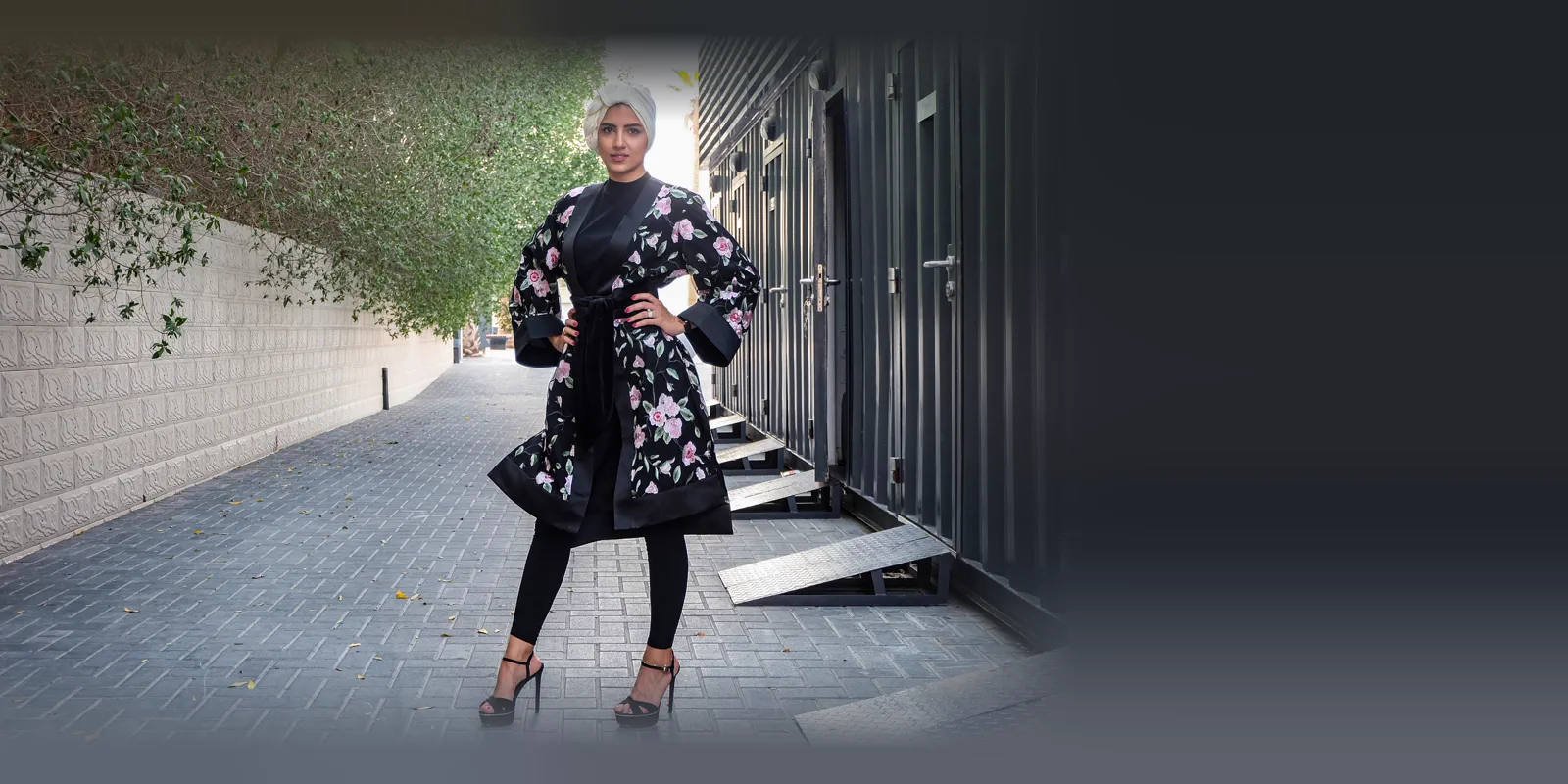 Woman in a knee-length black open abaya with pink floral embroidery over black trousers, standing in a paved lane under a tree canopy