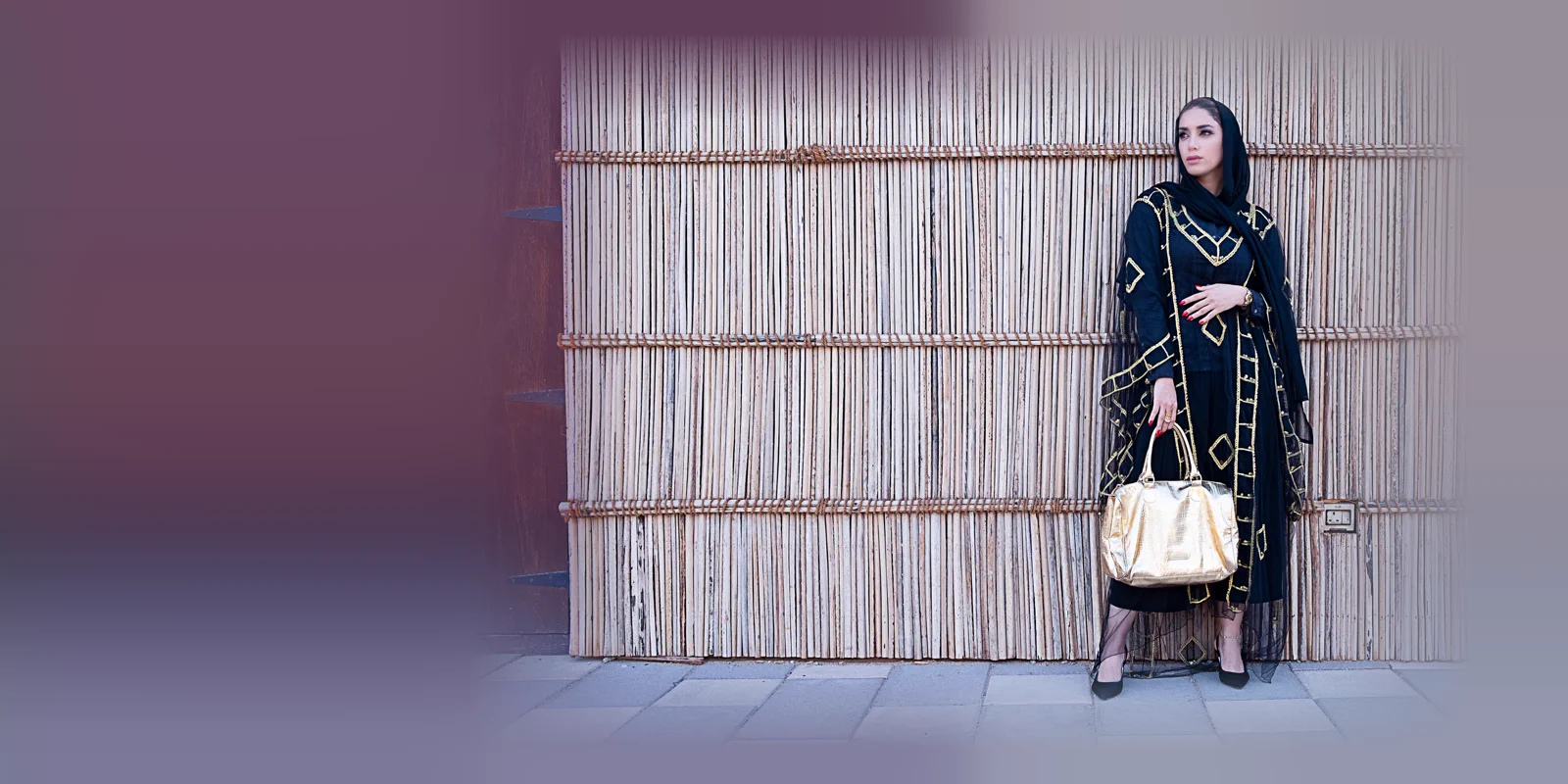 Woman in a black abaya with gold geometric outline detail and a black hijab holding a gold handbag in front of a woven bamboo wall