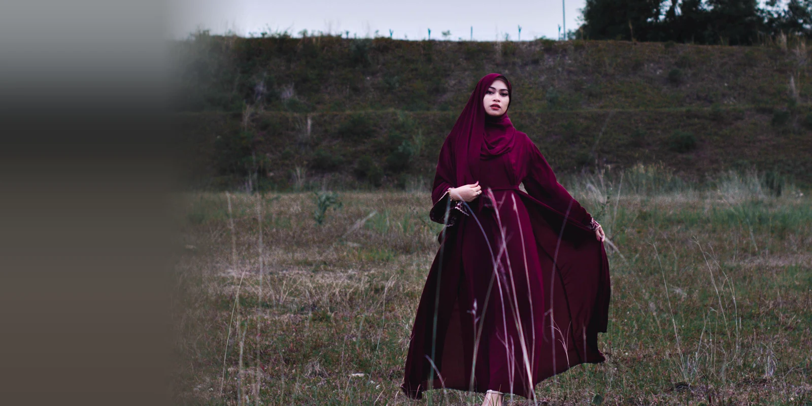 Woman in a burgundy abaya and headscarf standing in a grassy field under an overcast sky with the hem swept to one side