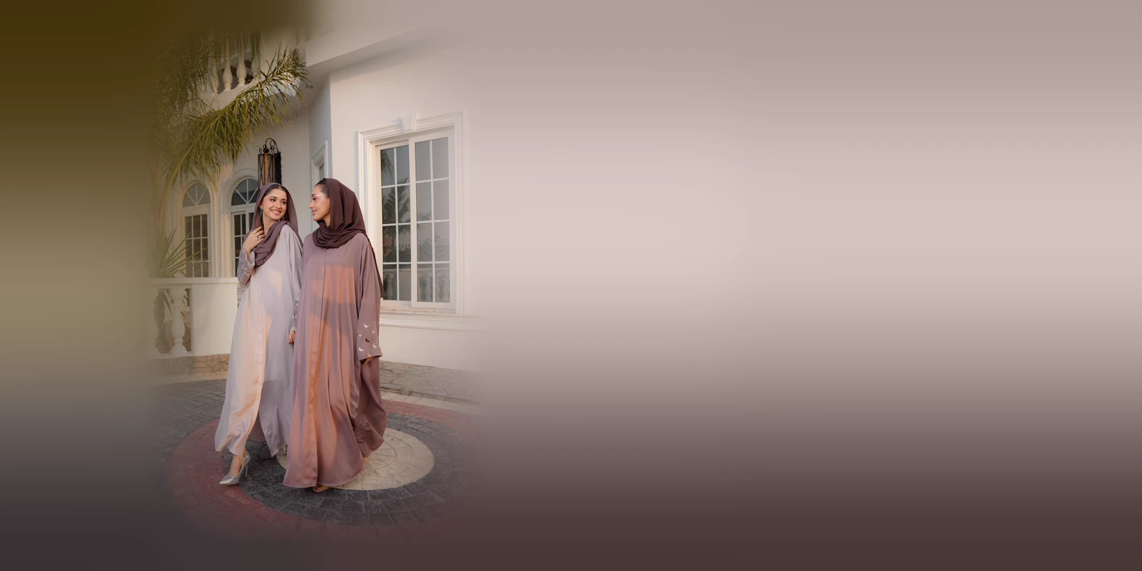 Two women in flowing abayas in soft grey and mauve with matching headscarves, smiling at each other in front of a white building