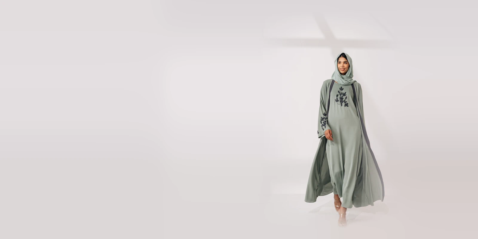 Woman in a sage green open abaya with dark floral embroidery and a matching hijab walking in a bright studio