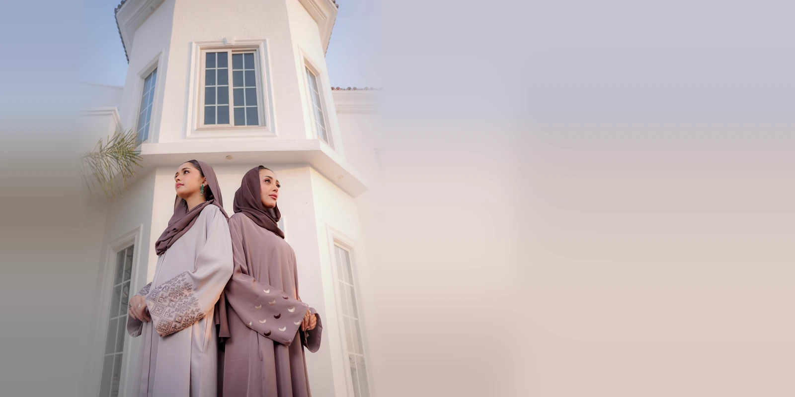 Two women in grey and mauve abayas with matching headscarves, seen from a low angle in front of a white tower