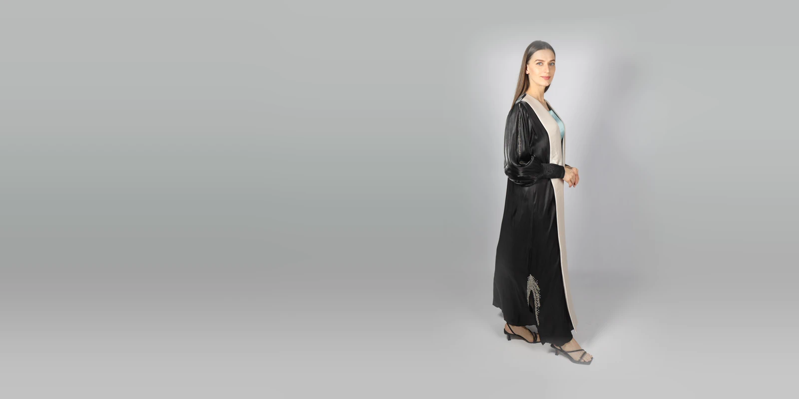 Woman in a black abaya with satin sleeves and a pale beige front panel, seen in profile against a grey studio backdrop