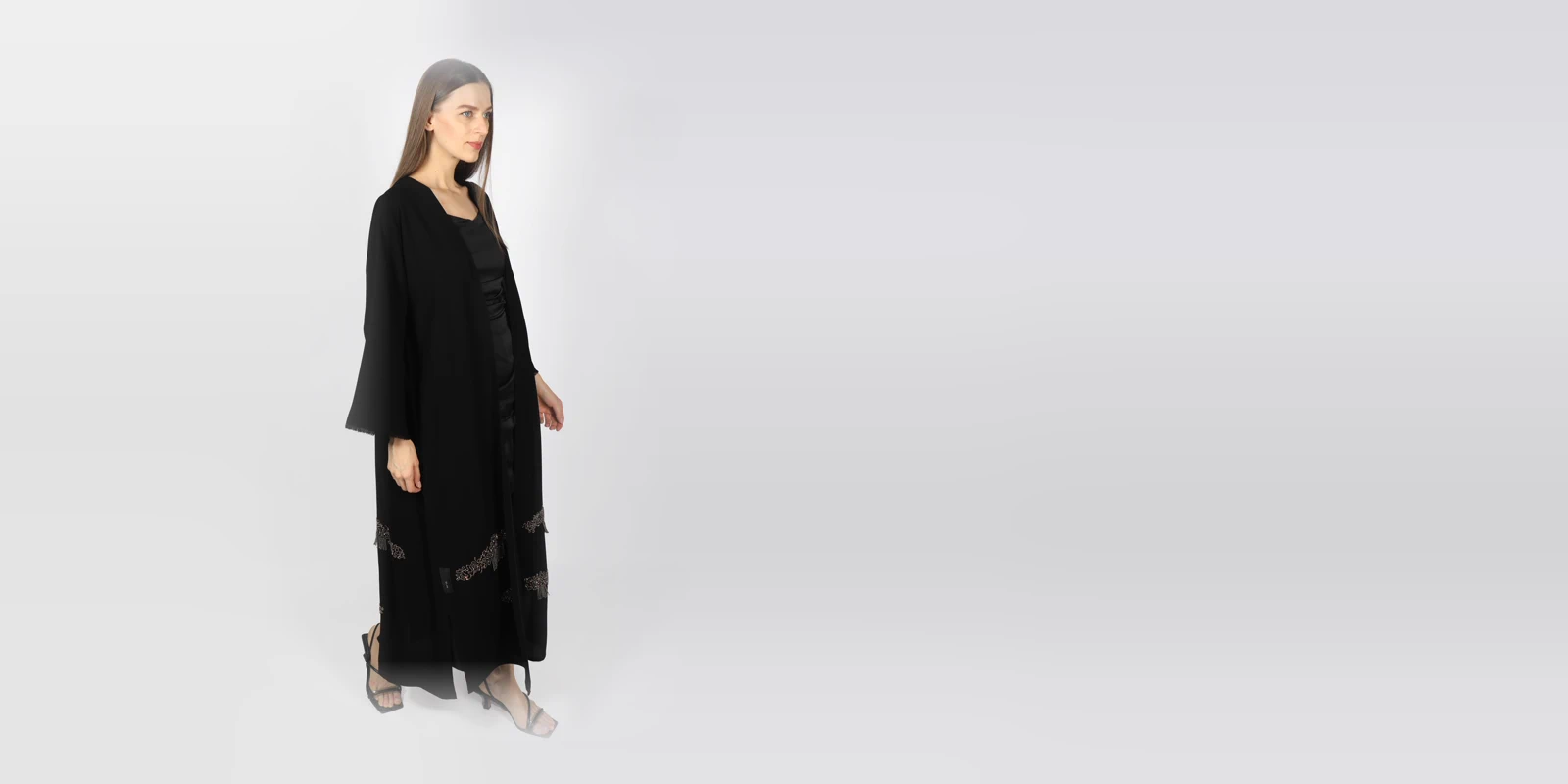 Woman in a long black open abaya over a black dress and strappy sandals, standing in three-quarter view against a grey studio backdrop