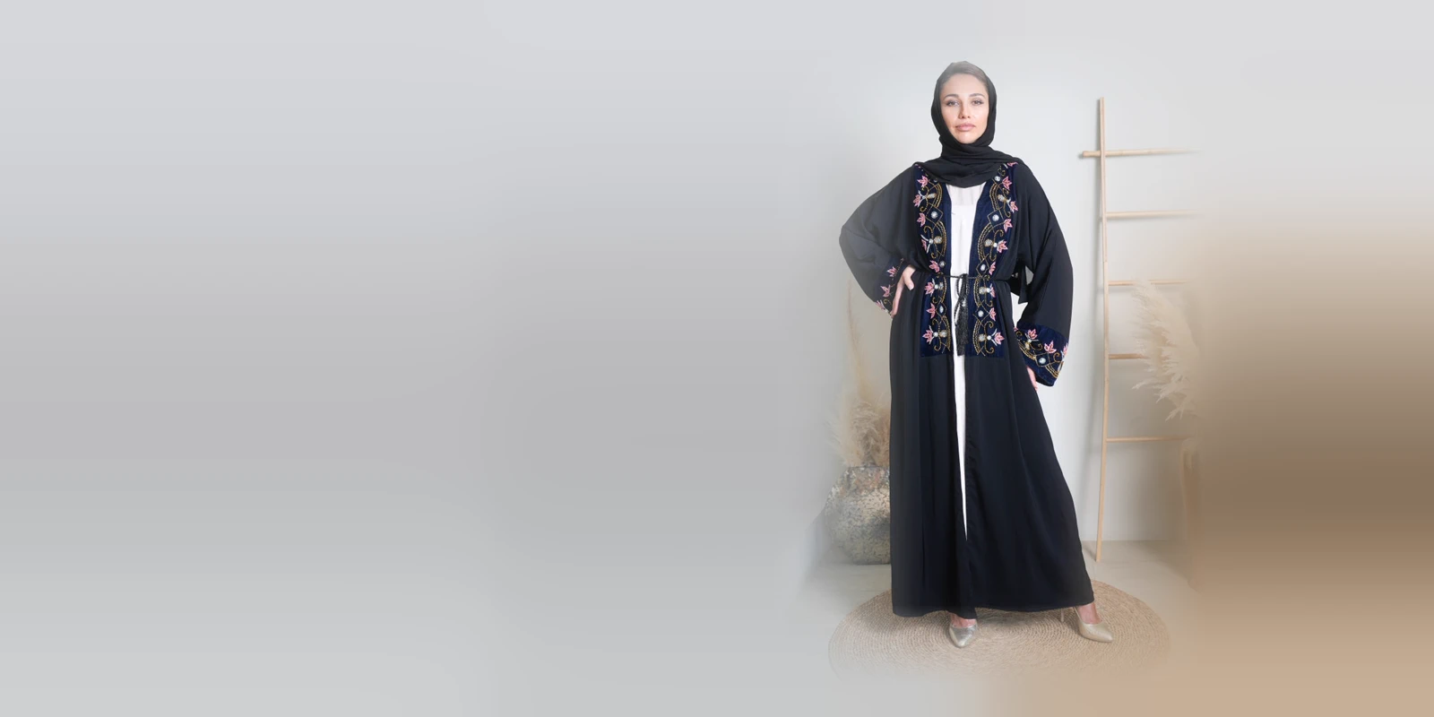 Woman in a black open abaya with floral embroidery on the front panel and a black headscarf, hand on hip, in a bright studio with a wooden ladder and dried pampas grass