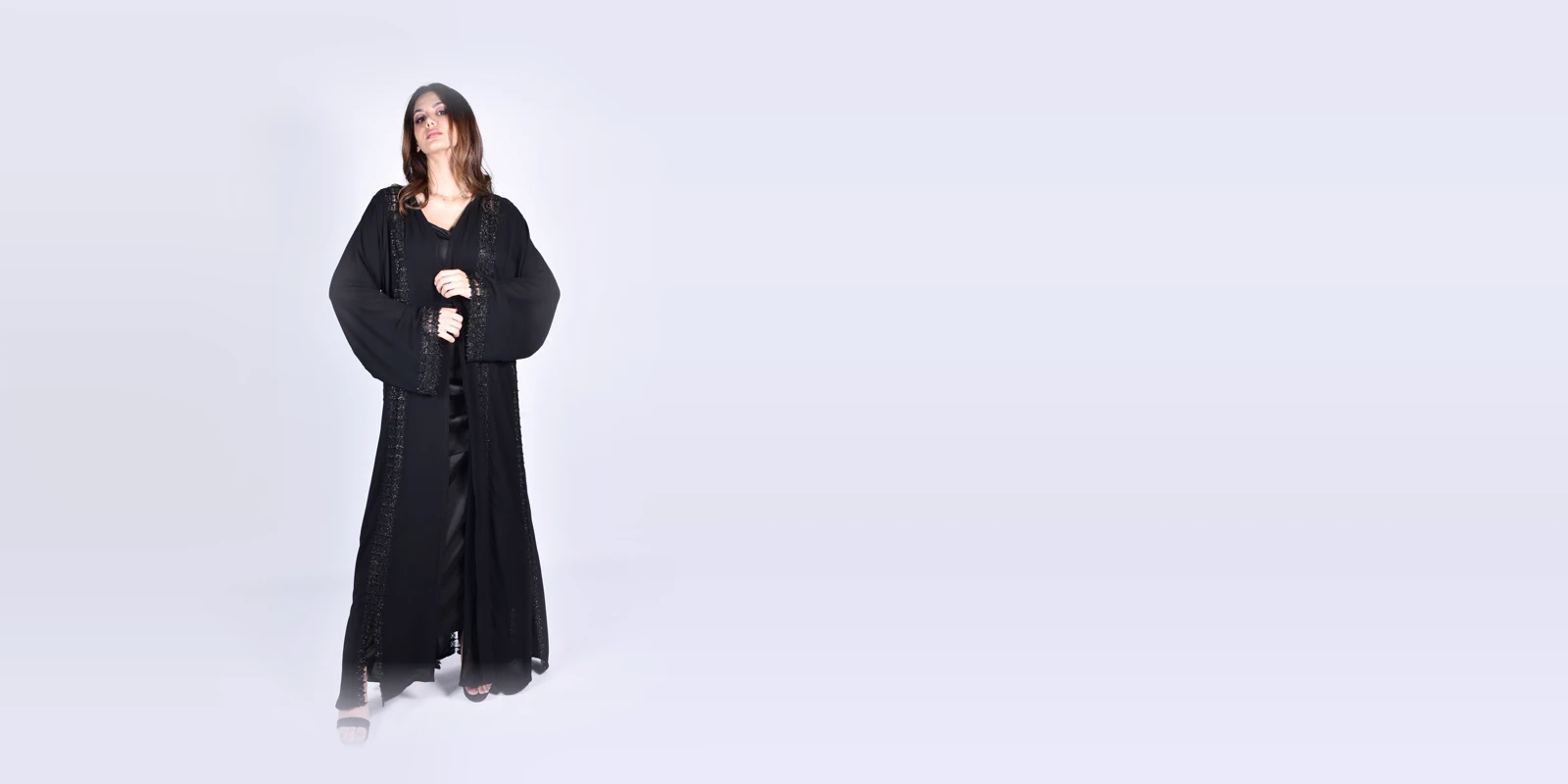 Woman in a black open abaya with lace trim over a black dress, hands together, against a light grey studio backdrop