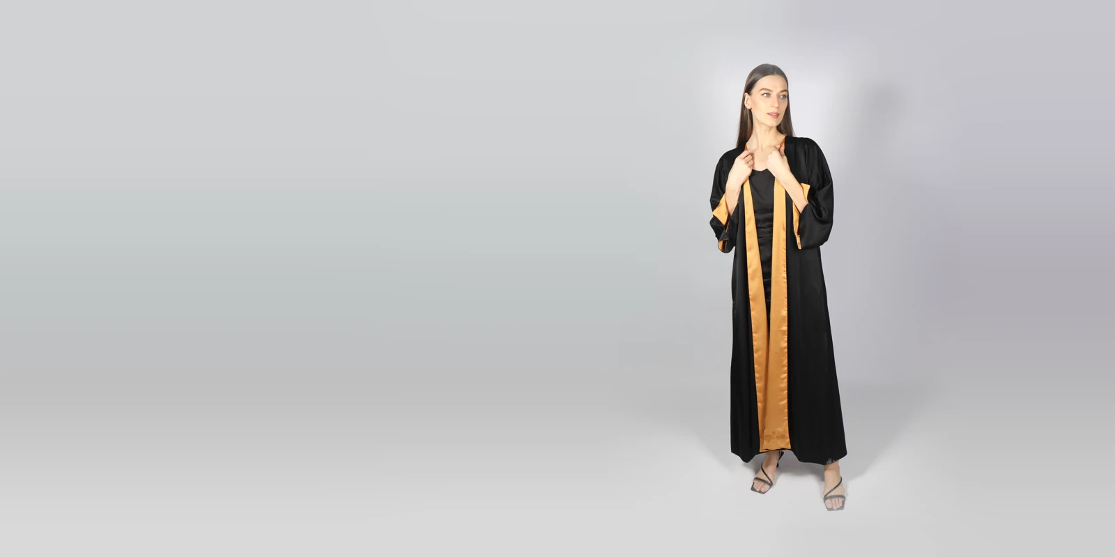 Woman in a black open abaya with golden satin lining, holding the front edges of the robe, against a light grey studio backdrop