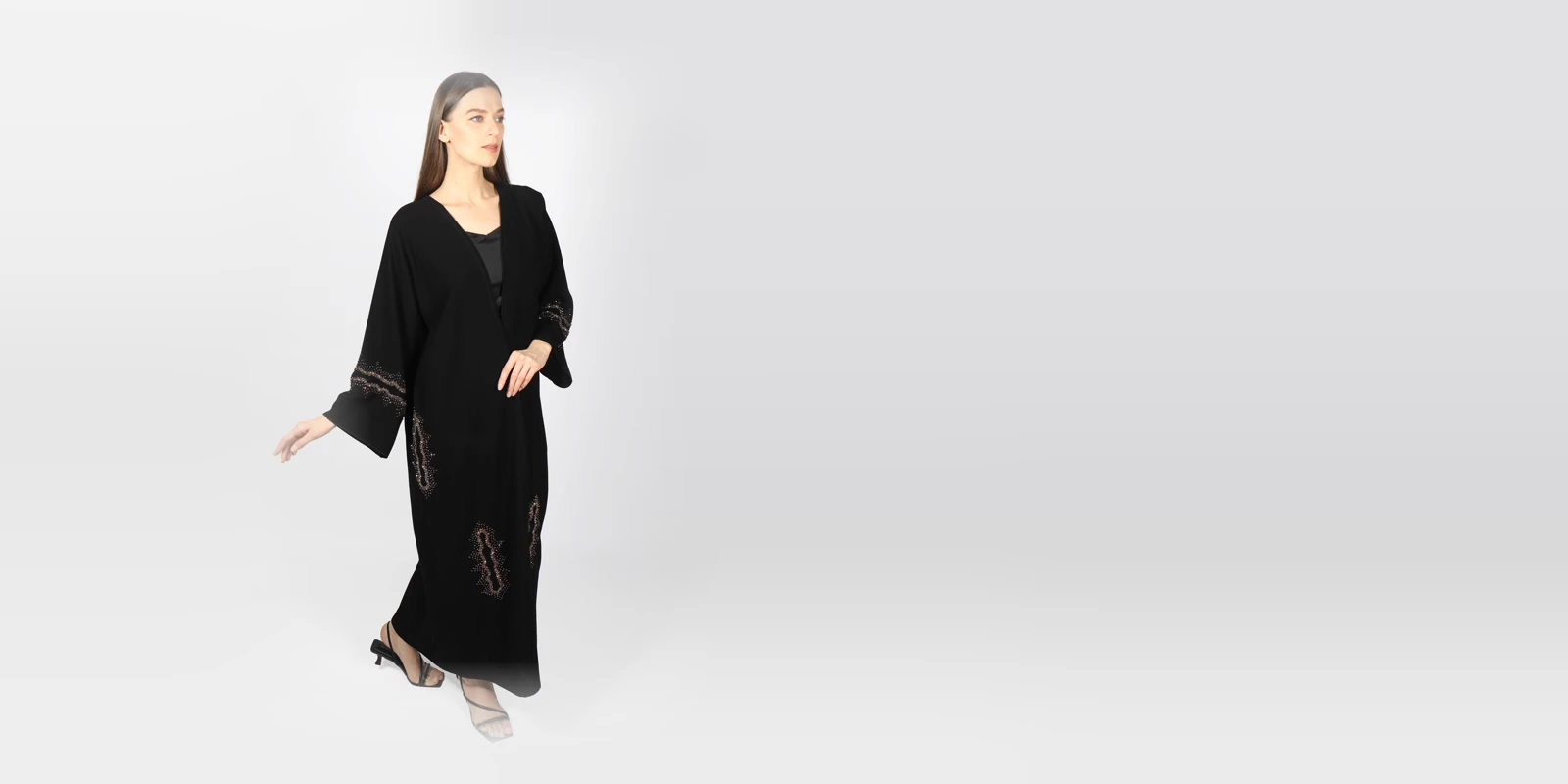 Woman in a long black open abaya with embroidered sleeves and details, walking against a light grey studio backdrop