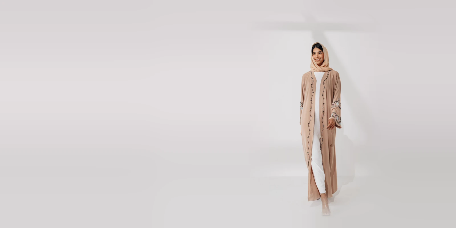 Woman in a tan open abaya with embroidered sleeves and a matching headscarf over a white dress, in a bright studio
