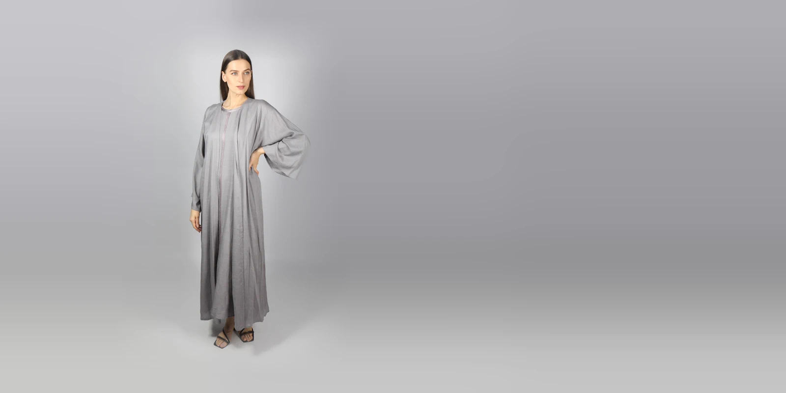 Woman in a loose grey abaya with wide sleeves, one hand on her hip, against a light grey studio backdrop