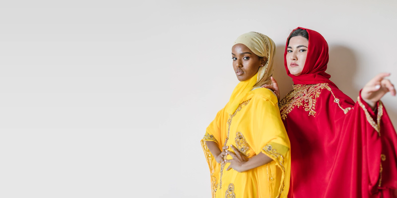 Two women in flowing embroidered kaftan-style abayas in yellow and bright red with matching headscarves, posing against a pale wall
